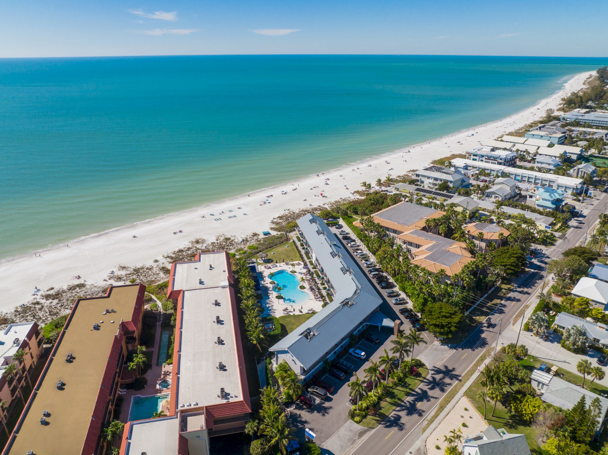 Aerial view of a coastal area with a sandy beach, turquoise water, beachfront buildings, and a pool.