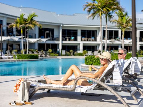 Two people lounge by a resort pool on sun chairs, woman in a hat sunbathing, man reading nearby, palm trees and a modern hotel backdrop.