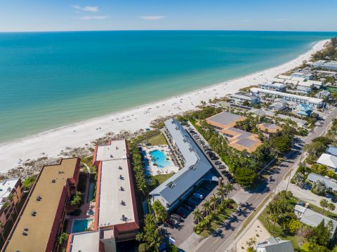 Aerial view of a coastal town with a white sandy beach, turquoise sea, palm-lined streets, and colorful buildings including a poolside complex.