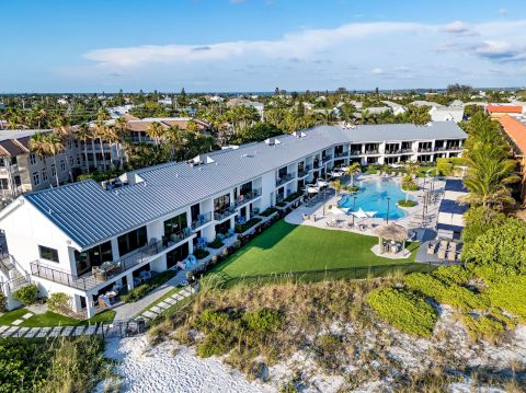 A beachfront resort features a large pool and lounge area, surrounded by lush greenery and sandy beach, with buildings in the background.