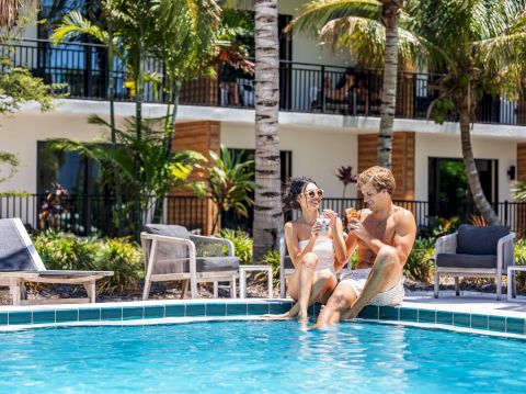 Two people sit by a resort pool, chatting and smiling as the sun shines on palm trees and lounge chairs nearby.