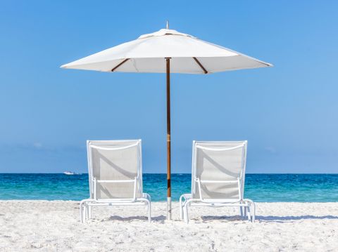 Two empty beach chairs under a white umbrella face the calm blue sea on a sunny sandy shore, inviting a relaxing seaside moment.