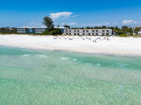 A sunny beach with clear turquoise water, white sand, and a row of low-rise buildings in the background under a blue sky.