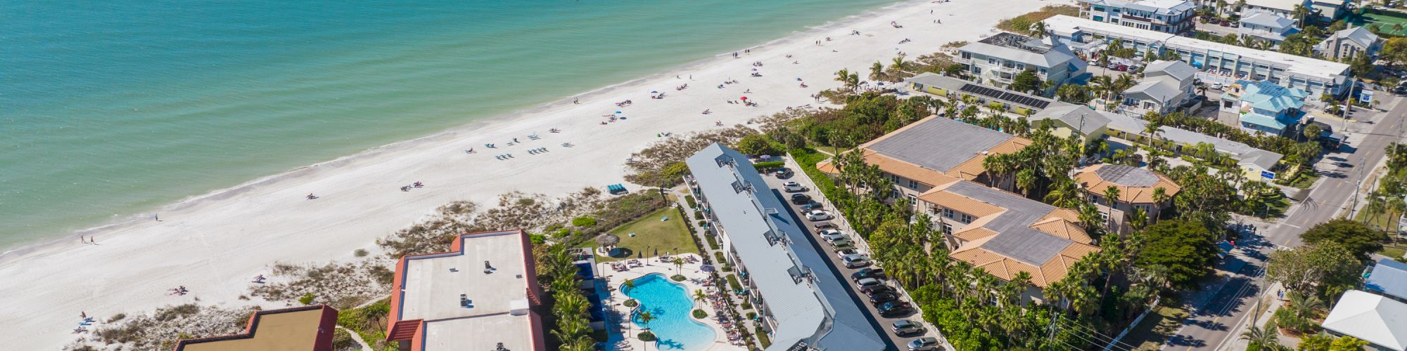 Aerial view of a coastal area with sandy beach, turquoise ocean, buildings, pools, and a road lined with houses and trees.