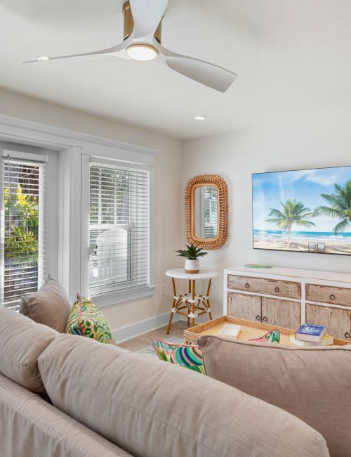 A cozy living room with a beige couch, white ceiling fan, tropical wall art, plants, and light streaming through a glass door with blinds.