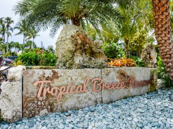 The image shows a sign for "Tropical Breeze Resort" surrounded by palm trees, rocks, and tropical plants in a landscaped setting.