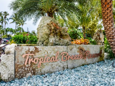 The image shows a sign for "Tropical Breeze Resort" surrounded by palm trees, rocks, and tropical plants in a landscaped setting.