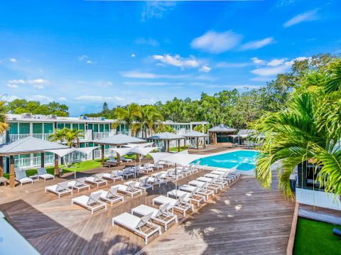 A resort-style pool area with multiple lounge chairs, shaded cabanas, palm trees, and a bright blue sky overlooking a calm pool.