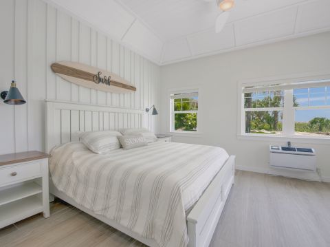 A bright coastal-style bedroom with a white paneled wall, a large bed, matching nightstands, blue lamps, and a view of greenery through a window.