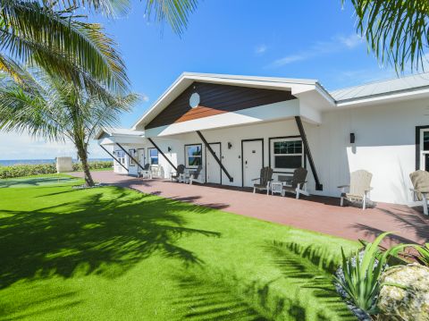 A sunny coastal home with a white facade, covered patio, palm trees, and a lush green lawn&mdash;perfect for outdoor lounging by the water.