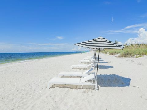 A sunny beach with white sand, calm blue water, and a row of empty lounge chairs under striped umbrellas facing the ocean.