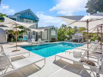 A serene poolside scene with loungers and umbrellas, set before a charming house surrounded by lush greenery and blue skies.