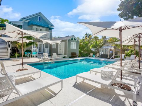 A serene poolside scene with loungers and umbrellas, set before a charming house surrounded by lush greenery and blue skies.