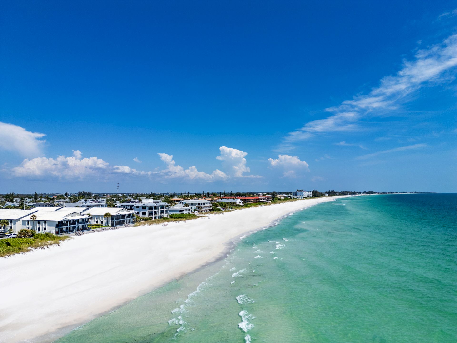 A sunny tropical beach with white sand, turquoise water, and houses lining the shore under a bright blue sky.