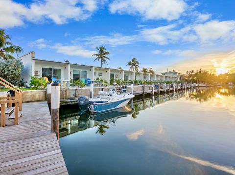 A waterfront view with a dock, a boat, palm trees, and houses under a blue sky and sunset.