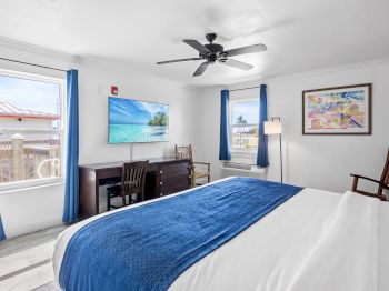 The image shows a bright bedroom with a bed, blue accents, a TV, desk, chair, and ceiling fan, creating a cozy, inviting atmosphere.
