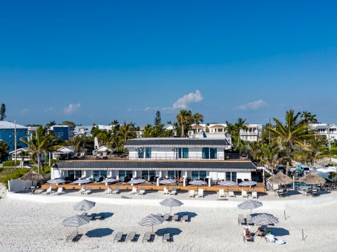 A sunny beachfront restaurant/hotel with white stone seating, palm trees, and numerous sun loungers under umbrellas on a sandy beach.