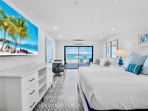 A bright, modern hotel room with a king bed, blue accents, a wall-mounted TV showing a tropical beach, and a balcony overlooking the ocean.