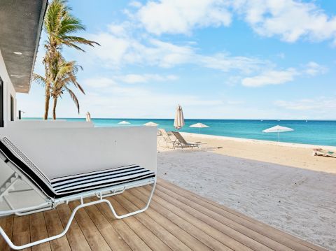 A sunny beach view with a wooden deck, white lounge chair, palm tree, and turquoise sea under a blue sky, inviting relaxation at the coast.