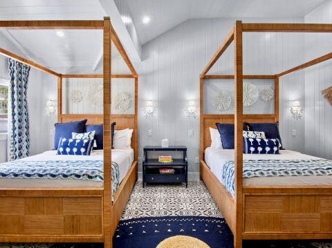 Two wooden four-poster beds sit opposite each other in a bright, airy bedroom with a central nightstand, blue-and-white linens, and a patterned rug, ending the sentence.