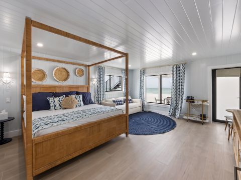 A bright coastal bedroom with a wooden four‑poster bed, blue and white decor, circular woven rug, windows with curtains, and a light, airy vibe. If you want a shorter, end-with-period version, I can trim it.
