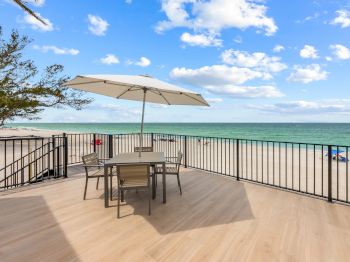 A beachside deck with a table, chairs, and umbrella overlooks the ocean under a blue sky with scattered clouds.