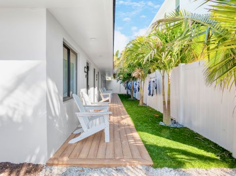 A bright hotel balcony with white chairs on a wooden deck, palm trees along a white wall, green lawn, and sunny blue sky.