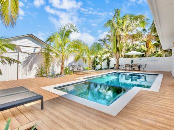 A modern backyard with a swimming pool, wooden deck, palm trees, lounge chairs, and an outdoor dining area under a blue sky.