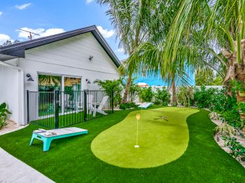 A backyard with a mini-golf area, surrounded by tropical plants and a white house on the side under a clear blue sky.