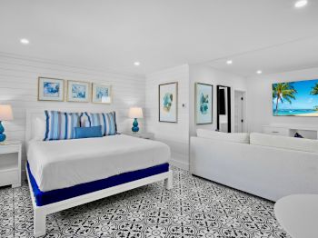 The image shows a bright bedroom with a bed, patterned floor, beach-themed decor, lamps, and ocean artwork on the wall.