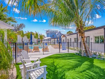 The image shows a sunny outdoor area with a pool, chairs, palm trees, and surrounding buildings, enclosed by a black fence.