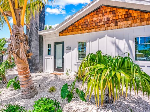 The image shows a house with a wooden facade, surrounded by tropical plants and a palm tree, under a blue sky with scattered clouds.