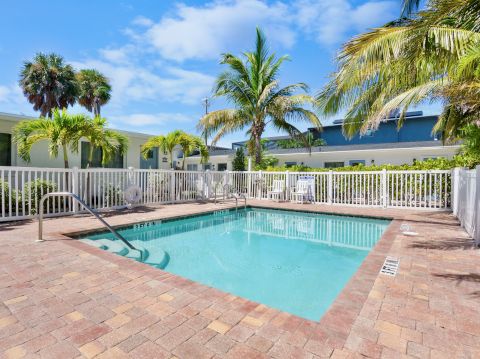 A rectangular community pool with clear blue water, white fence, palm trees, and lounge chairs in a sunny, tropical courtyard.