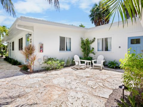 A sunny white cottage with a stone patio, two windows, blue door, and tropical plants; two white chairs sit by the entrance.