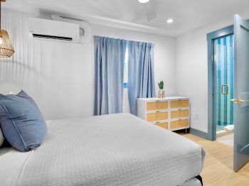 A cozy bedroom with a bed, blue curtains, and a chest of drawers near an open door leading to another room or space.