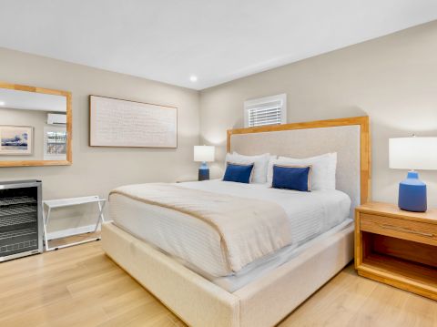A neatly arranged bedroom with a large bed, wooden furniture, two lamps, a mini-fridge, and wall art in a well-lit room.