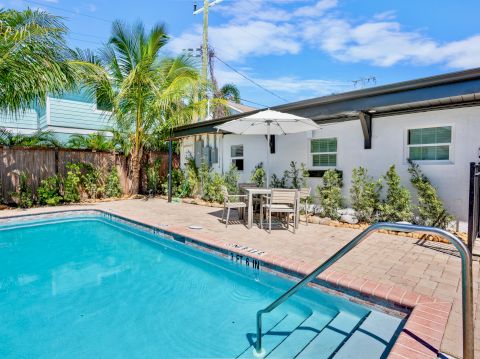 A sunny backyard with a small pool, palm trees, a white house, and a patio table under a large umbrella by the pool, bright and inviting.