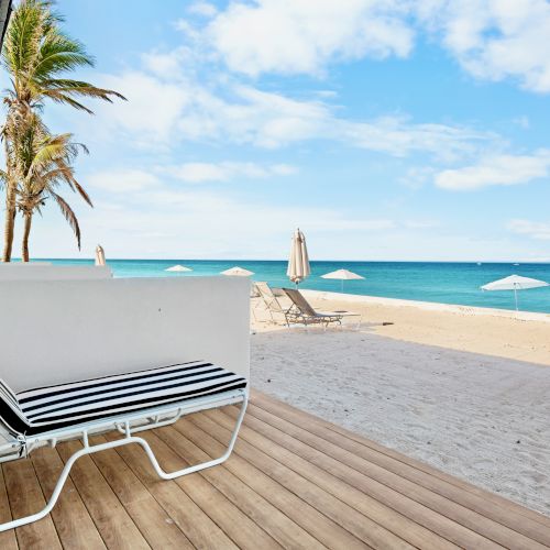 A beach view with a modern white lounger on a deck, palm trees, and a vast ocean under a blue sky with scattered clouds.