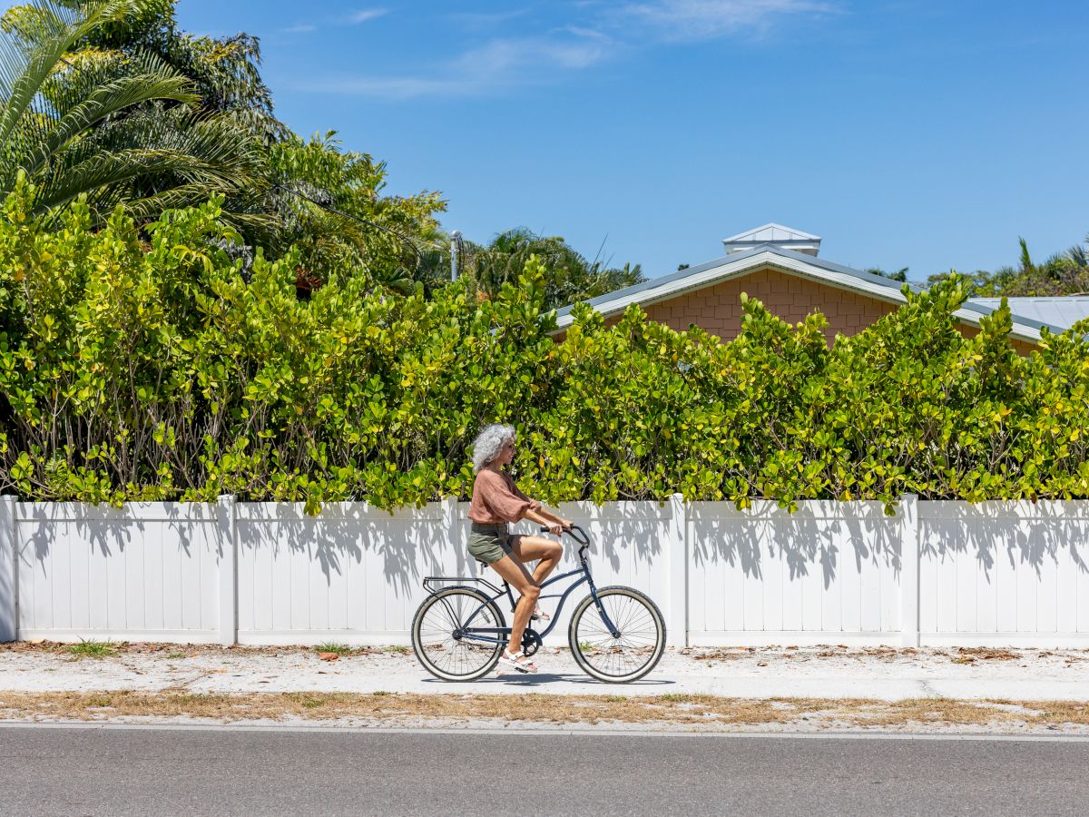 A person rides a bicycle on a sunny day past a white fence with greenery, under a clear blue sky.