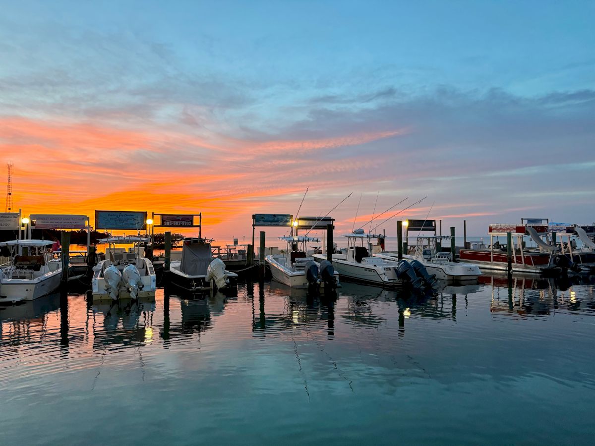 A serene marina at sunset with boats docked, calm water reflections, and a colorful sky transitioning from orange to blue.