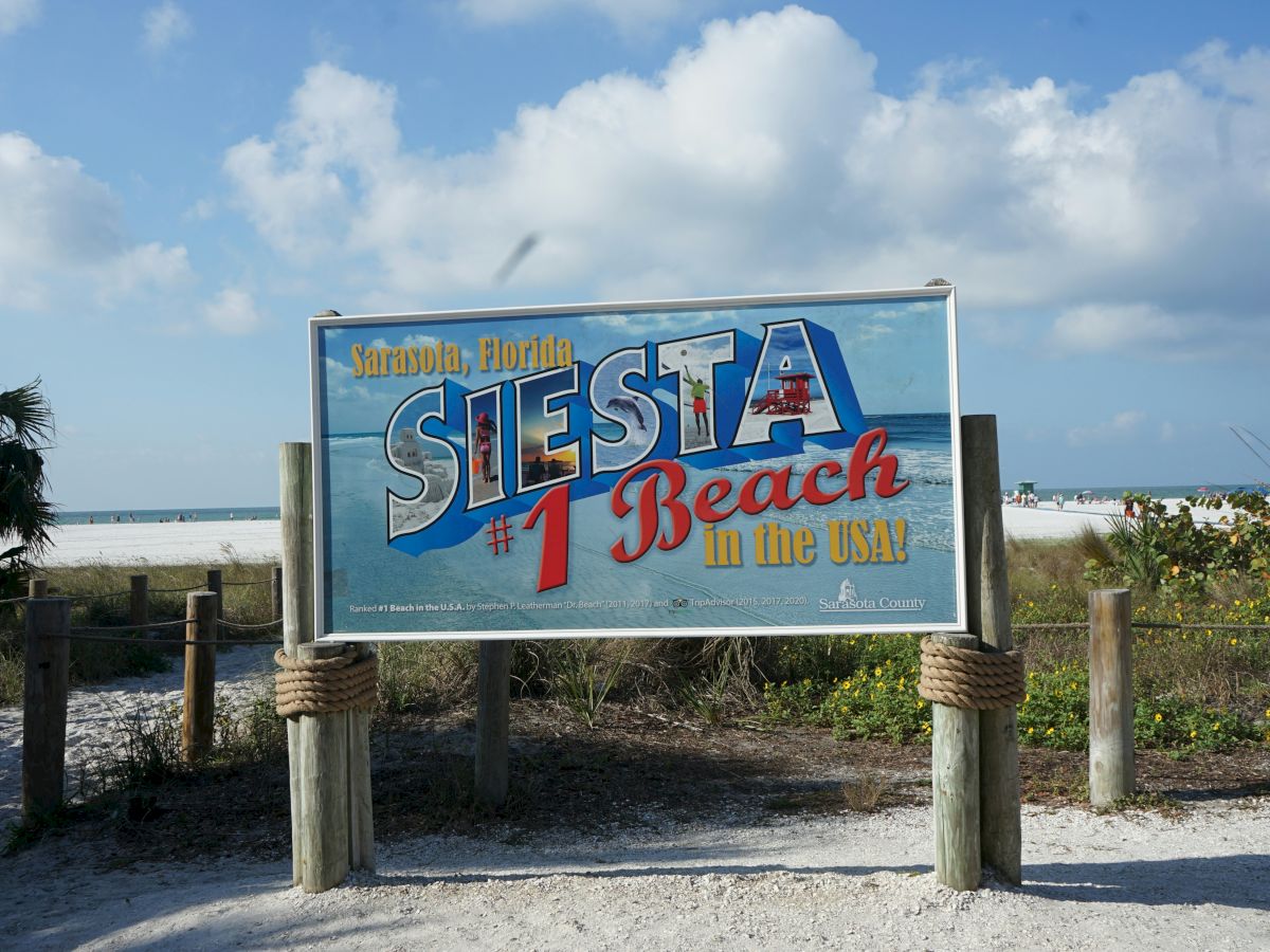 The image shows a sign for Siesta Beach with "No. 1 Beach in the USA" written on it, set against a beach and blue sky backdrop.