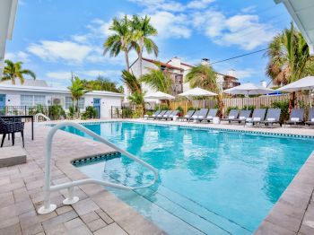 A sunny resort pool area with clear blue water, palm trees, loungers, umbrellas, and a bright sky.