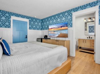 A bright hotel room with a blue patterned wallpaper, a king bed, blue door, wooden furniture, and a doorway to a bathroom.