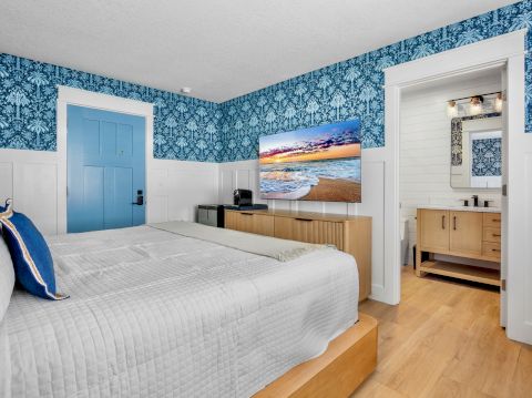 A bright hotel room with a blue patterned wallpaper, a king bed, blue door, wooden furniture, and a doorway to a bathroom.