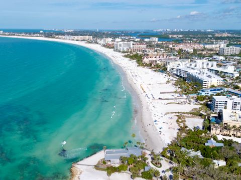 A vibrant coastal town with white-sand beaches, turquoise water, and modern beachfront buildings lining a curved coastline under a clear blue sky.