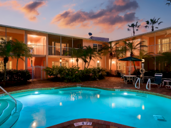 An outdoor hotel courtyard at sunset with a blue swimming pool, palm trees, lounge chairs, and a multi-story building surrounding the space.