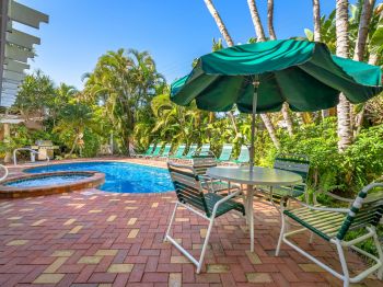 Tropical hotel pool with blue water, lounge chairs, and a green umbrella beside tall palm trees.