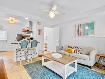 Bright open kitchen-living room with white walls, two blue chairs, couch, coffee table, ceiling fan, and framed artwork.